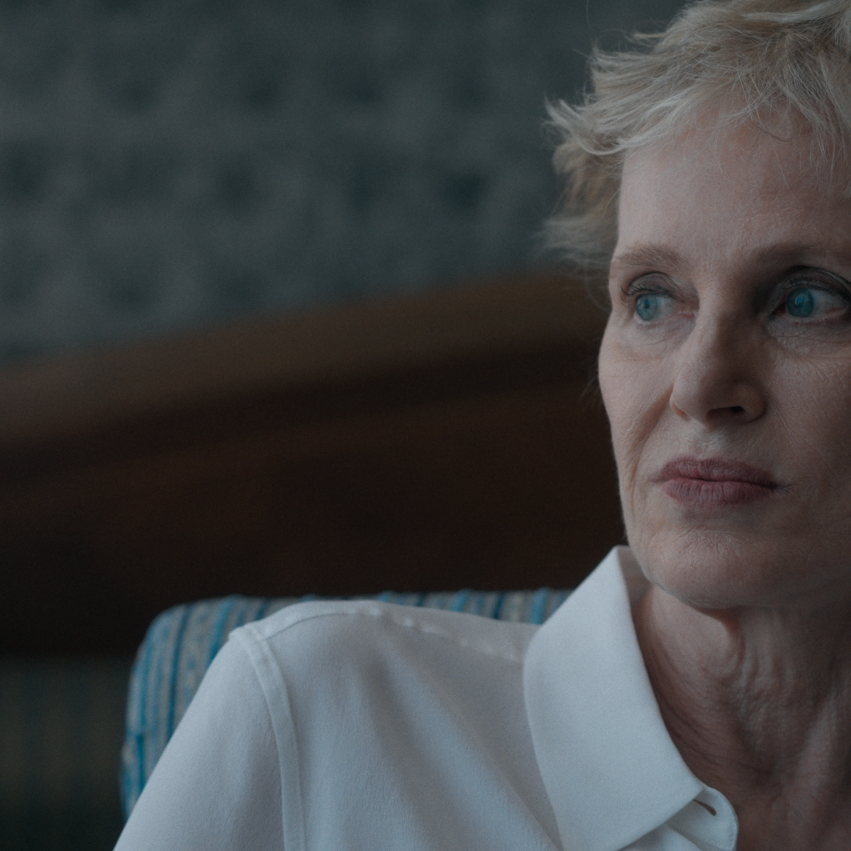 A lady in a white shirt sitting in a dark room, looking out at something. She has her hand behind her ears.