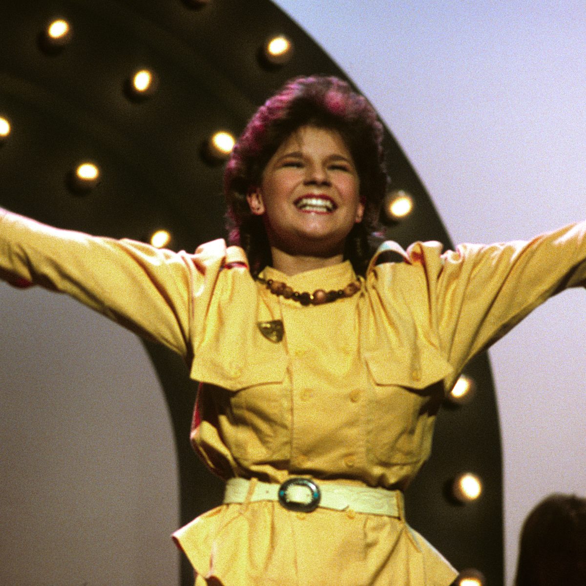 A young girl in yellow clothes hold her hands out while holding a microphone. She looks happy