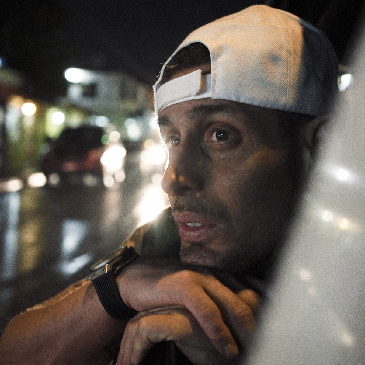 A guy with a cap hanging out the window of a car while driving through the city at night.
