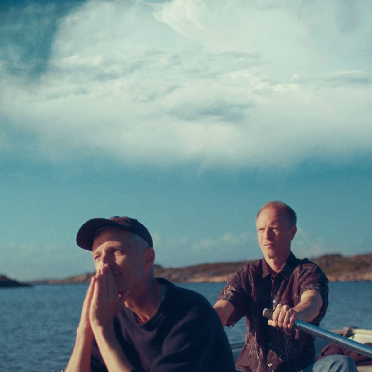 Two men sitting in a boat out at sea. One of them is rowing the boat and the other one has his hands tied before his face.