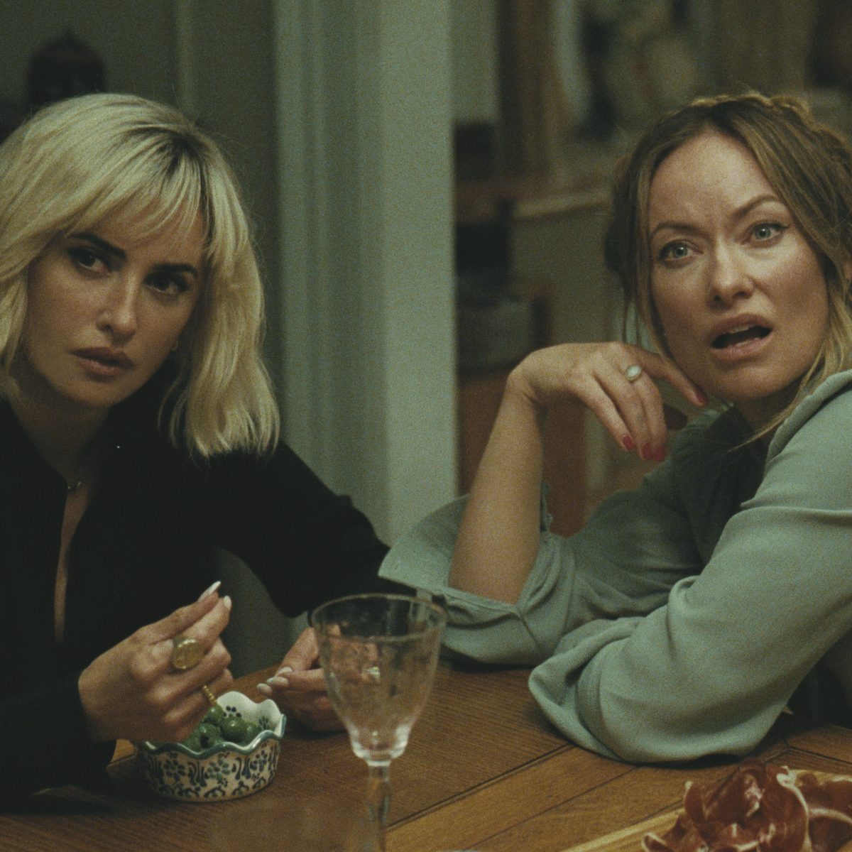 Two women leaned over a table. The woman to the right has brown hair and looks surprised. The woman to the left has blonde hair and has a neutral facial expression.