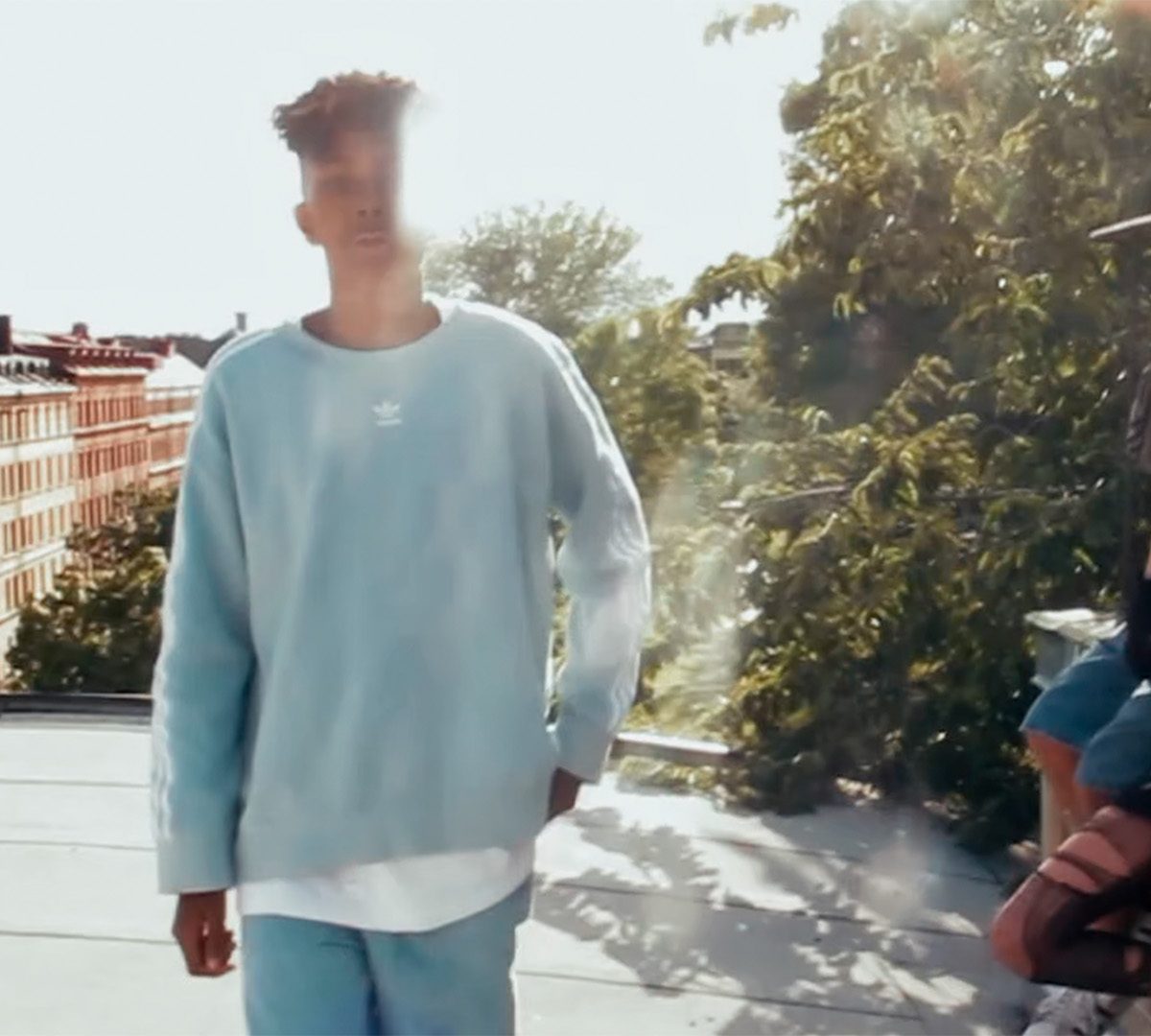 A young man wearing a blue sweatshirt with the sun against his back. Behind him are a boy and a girl, the boy is filming with a phone.